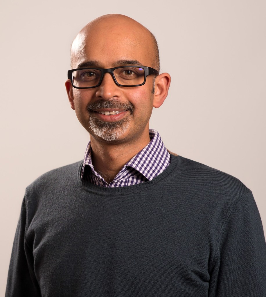 Nair has glasses and a short beard. He is facing the camera smiling wearing a purple checked shirt and a black jumper.