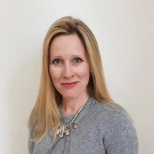 Natalie looking towards the camera smiling. She has long blond hair and blue eyes. Natalie is wearing a light grey top and necklace.
