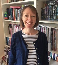 Rebecca has short brown hair in a bob and brown eyes. She is standing in front of a bookcase smiling wearing a blue and white stripy top and navy cardigan.