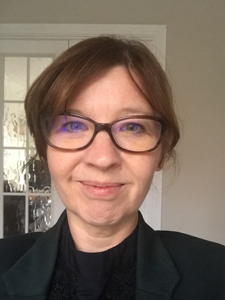 Melanie has blue eyes and a brown fringe. She is wearing glasses, looking at the camera smiling and wearing a black blouse.